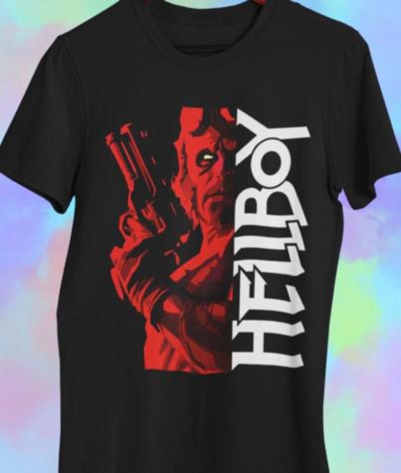 Puede incluir: Camiseta negra con un gr&aacute;fico rojo de un personaje sosteniendo un arma, con la palabra "HELLBOY" en texto vertical blanco. El dise&ntilde;o es una ilustraci&oacute;n audaz de estilo c&oacute;mic.