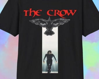 May include: Black t-shirt featuring the text "THE CROW" in red above a graphic of a crow with open eyes. Below the crow is a vertical white rectangle with a person in black clothing walking towards the viewer.