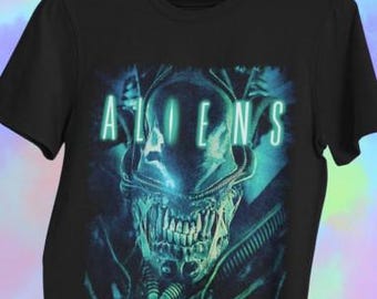 May include: Black t-shirt featuring a detailed, teal and blue graphic of an alien creature's head. The word "ALIENS" is displayed in teal block letters above the creature. The design is centered on the shirt.
