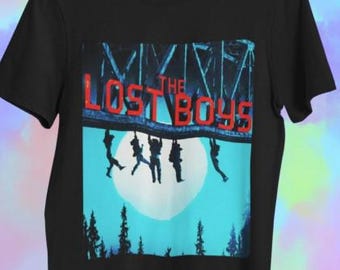 Op de afbeelding: Zwart t-shirt met een grafisch ontwerp van "The Lost Boys". Het ontwerp bevat de tekst "THE LOST BOYS" in het rood, met silhouetten van figuren die aan een brug hangen tegen een blauwe lucht en volle maan.