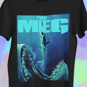 May include: Black t-shirt featuring a teal graphic from the movie "The Meg." The design shows a shark leaping from the ocean, with the word "THE MEG" in large teal letters above. The image has a blue ocean background.