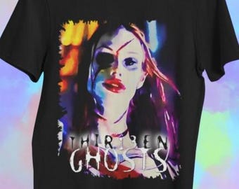 Puede incluir: Camiseta negra con un retrato estilizado de una mujer con una herida en la cara y pintalabios rojo. El texto "THIRTEEN GHOSTS" está impreso en blanco. La camiseta está colgada en una percha de madera.