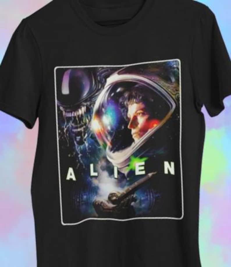 May include: Black t-shirt with a graphic from the film "Alien." The design features a person in a spacesuit, the film's title, and an alien creature. The image is framed in white.