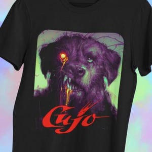May include: Black t-shirt featuring a graphic of a snarling dog with a glowing red eye and dripping yellow liquid. The word "Cujo" is written in red, stylized lettering below the dog's face.