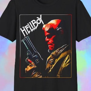 May include: Black t-shirt with a graphic of Hellboy, a red demon with horns, holding a gun. The word "HELLBOY" is printed in white above the character. The graphic is framed with a red and white border.