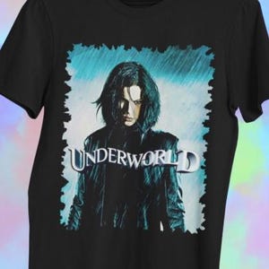 Puede incluir: Camiseta negra con un gráfico de la película "Underworld". El diseño presenta a una persona con cabello oscuro y una chaqueta de cuero negra, sobre un fondo azul y blanco. La palabra "UNDERWORLD" está impresa en letras plateadas.