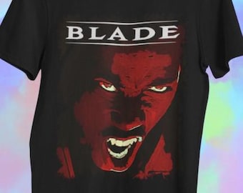 May include: Black t-shirt with a red graphic of a face with an angry expression and fangs. The word "BLADE" is printed in white block letters above the face. The shirt is made of a soft material.