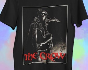 May include: Black t-shirt with a graphic from the film "The Crow." The design, within a white border, shows a figure with a crow and the title in red. The shirt has a crew neck.