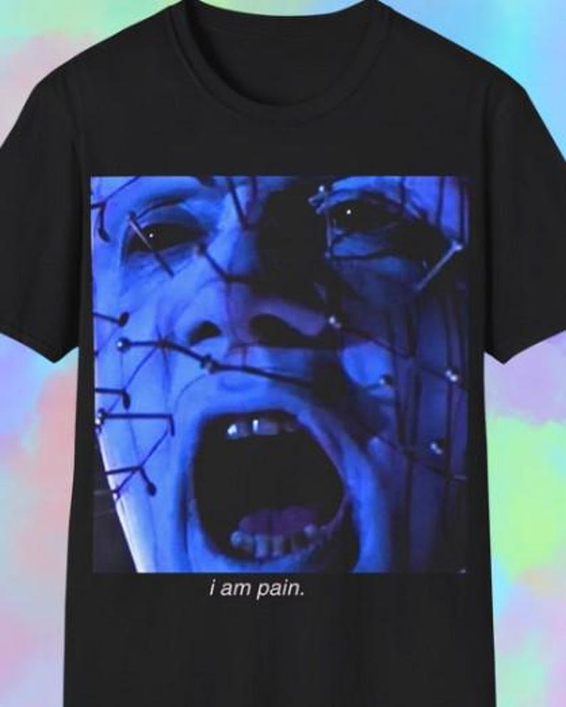 May include: Black t-shirt with a blue-toned face graphic, pins, and an open mouth. The text "i am pain" is printed below. The shirt has a crew neck design, a common style for casual wear.