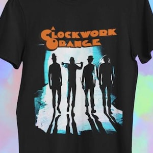Op de afbeelding: Zwart t-shirt met een afbeelding uit de film "A Clockwork Orange". Het ontwerp toont de titel van de film in oranje en vier silhouetten van figuren in hoeden en pakken, tegen een blauw-witte achtergrond.