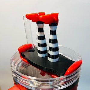 May include: Red travel mug with a clear lid and a whimsical topper featuring a pair of witch legs. The legs are black and white striped with bright red shoes. The mug has a red handle and accents.