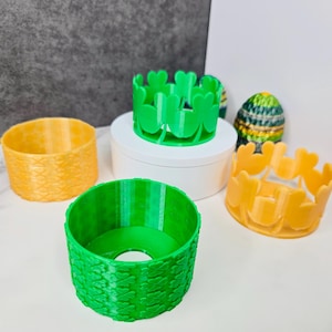 May include: A collection of 3D-printed decorative rings in various colors. The rings include a textured orange ring, a green ring with a woven pattern, a green ring with clover cutouts, and a yellow ring with heart-shaped cutouts. A decorative egg is also visible.