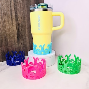 May include: A yellow Stanley mug with a handle, resting on a blue Easter-themed coaster. Three additional coasters in pink, green, and blue are displayed. The coasters feature a bunny and egg design, perfect for spring.