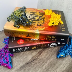May include: Two books titled "Iron Flame" and "Fourth Wing" with dragon-shaped bookmarks. The bookmarks are in various colours: yellow, blue, purple, and green. The books are stacked on a wooden surface.