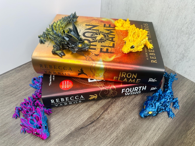 May include: Two books titled "Iron Flame" and "Fourth Wing" with dragon-shaped bookmarks. The bookmarks are in various colours: yellow, blue, purple, and green. The books are stacked on a wooden surface.
