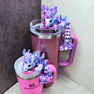 May include: Two Stanley tumblers, one pink and one purple, each adorned with a decorative dragon. The dragons are iridescent with purple and blue hues. The tumblers have handles and straws, perfect for cold beverages.