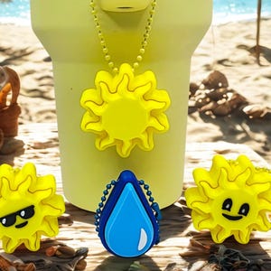 May include: A yellow water bottle with a sun-shaped charm necklace. Two additional sun-shaped charms with sunglasses and a smiling face are also present. A blue water droplet charm is also visible. The items are on a wooden surface, with a beach scene in the background.