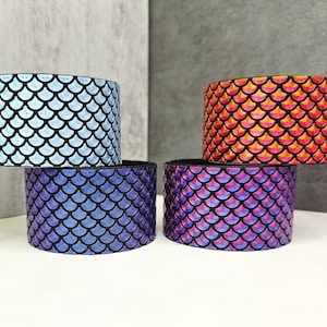 May include: Four cylindrical bracelets with a mermaid scale pattern. The bracelets are in shades of blue, purple, and orange. The scales are outlined in black, creating a textured appearance. The bracelets are stacked on top of each other.