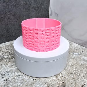 May include: A bright pink, cylindrical object with a repeating heart pattern. The object is sitting on a white, circular platform. The background is a mix of gray and white.