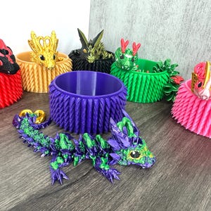 May include: A selection of vibrant 3D-printed dragon ornaments and coordinating decorative pots. The dragons are in shades of purple, green, gold, red, and pink. The pots are cylindrical with textured sides.
