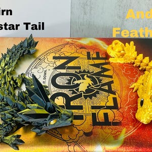 May include: Two dragon tail figurines, one green and black, the other gold, rest on a book. The green dragon is labeled "Tairn Morningstar Tail." The gold dragon is labeled "Andarna Feather Tail." The book cover is red and gold.