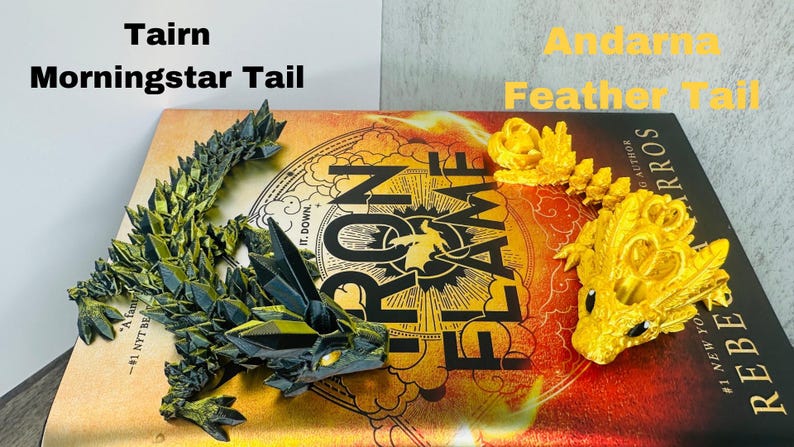 May include: Two dragon tail figurines, one green and black, the other gold, rest on a book. The green dragon is labeled "Tairn Morningstar Tail." The gold dragon is labeled "Andarna Feather Tail." The book cover is red and gold.
