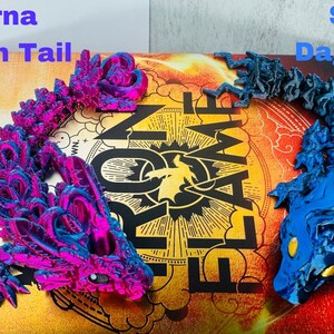 May include: Two 3D-printed dragon tail models, one magenta and blue, labelled "Andarna Scorpion Tail," and the other blue, labelled "Sgaeyl Dagger Tail." They rest on a book with the title "Flame and Shadow."