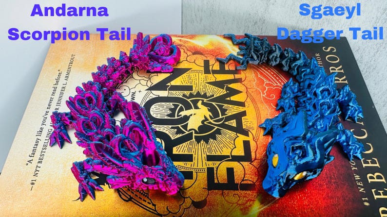 May include: Two 3D-printed dragon tail models, one magenta and blue, labelled "Andarna Scorpion Tail," and the other blue, labelled "Sgaeyl Dagger Tail." They rest on a book with the title "Flame and Shadow."