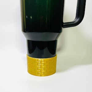 May include: A dark green and black travel mug with a black handle and a gold-colored brick-patterned base. The mug is designed to keep beverages hot or cold. The mug is made of stainless steel.