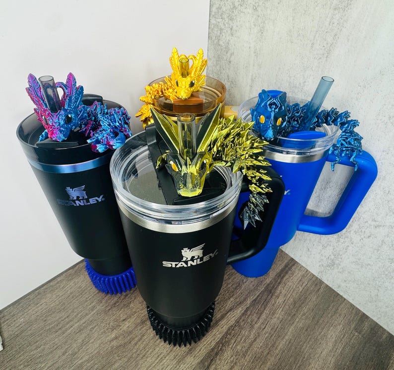 May include: Three Stanley travel mugs with dragon-themed lids. One black mug has a blue and purple dragon, another black mug has a gold dragon, and a blue mug has a blue dragon. Each mug has a handle and a lid.