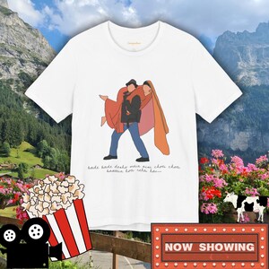 Puede incluir: Camiseta blanca con una ilustración de dibujos animados de una pareja bailando. El texto "bade bade deshko mera aya chotu chotu baatein hoti rehti hai..." está impreso debajo de la ilustración. La camiseta es un diseño con temática de cine.