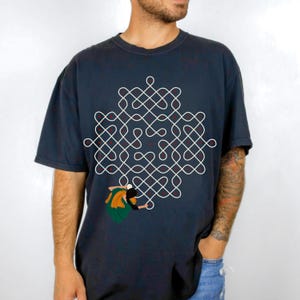 May include: Dark navy blue t-shirt featuring a white geometric design with red accents. The shirt also has a graphic of a person wearing green and orange clothing. The t-shirt is made of a soft material.