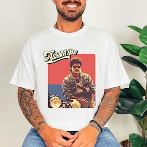May include: White t-shirt featuring a retro graphic of a man wearing sunglasses and a jacket, seated on a motorcycle. The graphic includes the text "Awara Hoon" in a stylized font, set against a red and blue background. The shirt is a casual wear item.