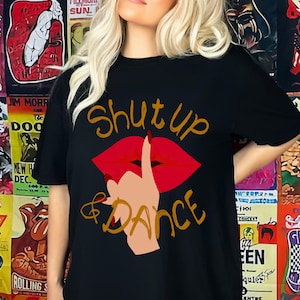 May include: Black t-shirt with a graphic design featuring red lips, a hand with a finger to the lips, and the text "Shut up & Dance" in gold. The shirt is a casual style.