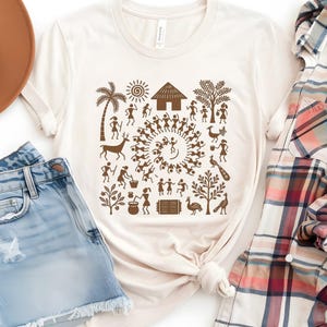 May include: A beige t-shirt with a brown graphic print of a tribal scene. The design features a circle of people dancing, a hut, trees, animals, and other symbols.