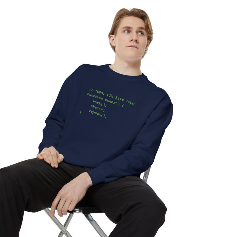 A blond man wearing a dark blue crewneck sweatshirt with green programming text and dark pants, seated on a metal chair.