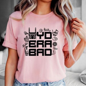May include: A light pink t-shirt with a black graphic design. The design includes the text "HYDERABAD" in a bold font, along with various illustrations of cultural symbols and food items. The shirt has short sleeves and a crew neck.