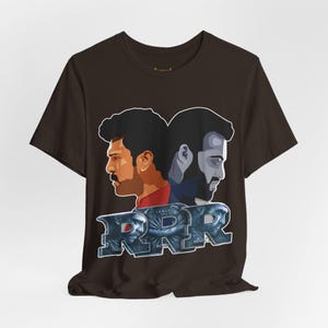 May include: A brown t-shirt featuring a graphic of two men's profiles and the text "RRR" in a stylized font. The graphic is in shades of red, blue, and gray. The t-shirt is made of a soft material.