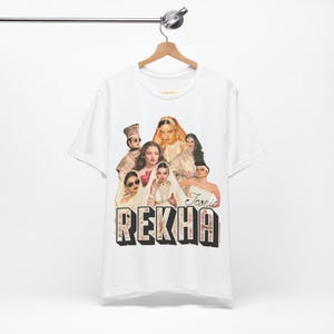 May include: White t-shirt featuring a graphic of several women in ornate attire, with the word "REKHA" in large, stylized letters. The word "Iconic" is also visible. The shirt is hanging on a hanger.