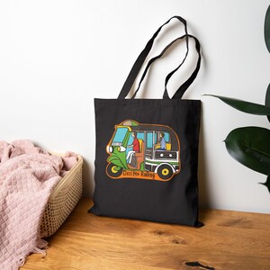 May include: Black tote bag featuring a colorful illustration of a green and yellow auto-rickshaw with passengers. The text "Desi Me Rolling" is below the vehicle. The bag has black handles and is made of a durable fabric.