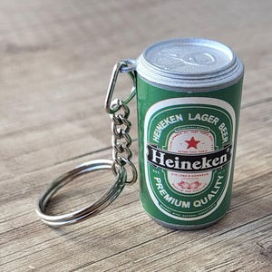 May include: A green Heineken Lager Beer can keychain with silver accents. The keychain features the Heineken logo and text on the can. The keychain is attached to a silver chain and ring.