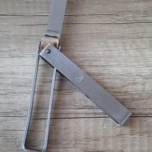 Könnte beinhalten: Ein silberfarbenes Klappmesser aus Metall mit einem rechteckigen Griff, der die geprägten Buchstaben LB aufweist. Das Messer hat eine einzelne gerade Klinge, die nach oben ausgeklappt ist, und eine dünne, rechteckige Metallschlaufe am Gelenk.