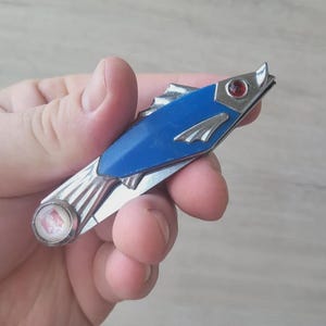 以下が含まれることがあります： 魚の形をしたヴィンテージのポケットナイフ。青いボディ、シルバーのアクセント、赤い宝石の目があります。ナイフは手で持たれています。