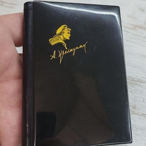 May include: A small, black book with a gold illustration of a person in a hat and a signature. The book has a glossy finish and a dark spine. The cover features the illustration and text in gold.