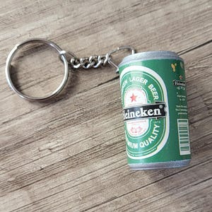 May include: A green and white Heineken beer can keychain with a silver keyring and chain. The can features the Heineken logo and text, with a red star. The keychain is on a wooden surface.
