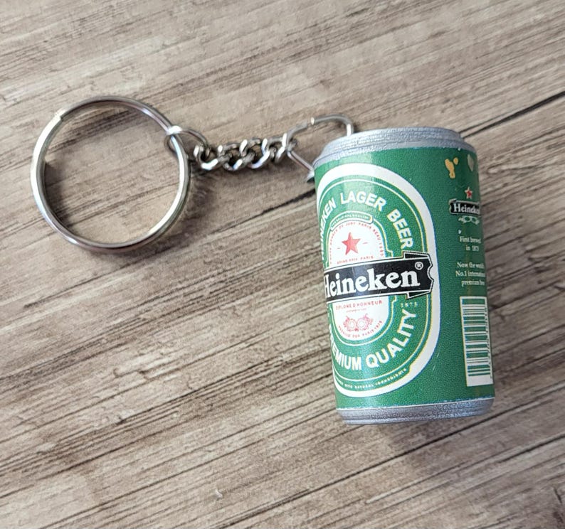 May include: A green and white Heineken beer can keychain with a silver keyring and chain. The can features the Heineken logo and text, with a red star. The keychain is on a wooden surface.