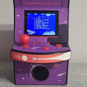 Peut inclure: Mini borne d'arcade violette miniature avec un joystick et des boutons rouges. L'écran affiche une liste de jeux, dont "F-22" et "Racing Fighter". Le texte "Experiment as you need" est au-dessus de l'écran. Le meuble a un haut-parleur et un design spatial.