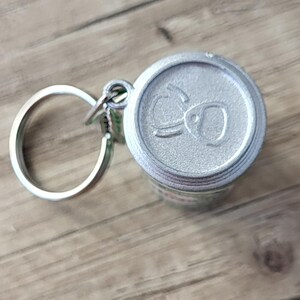May include: A silver-colored keychain featuring a miniature soda can design. The can top has embossed details. The keychain includes a silver ring and clasp. The background is a light brown wooden surface.