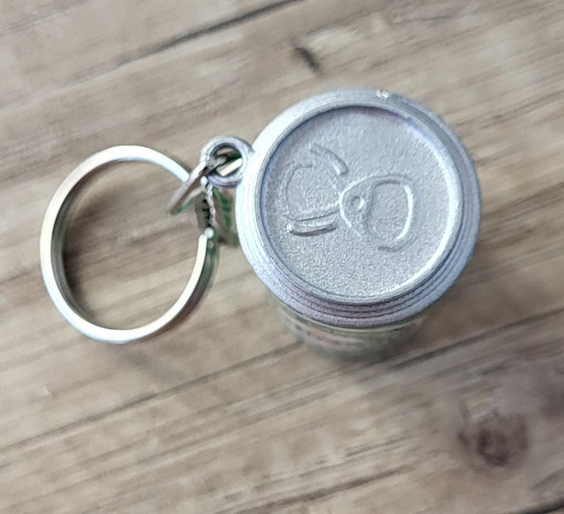 May include: A silver-colored keychain featuring a miniature soda can design. The can top has embossed details. The keychain includes a silver ring and clasp. The background is a light brown wooden surface.