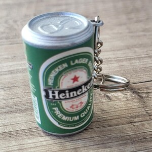 May include: Miniature green and silver Heineken Lager Beer can keychain. The can features the Heineken logo, a red star, and the words "PREMIUM QUALITY". A silver chain and keyring are attached.